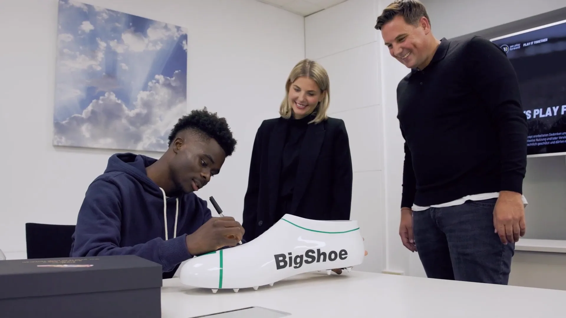 Bukayo Saka signing a white shoe with BigShoe written on it, a woman and a man looking on